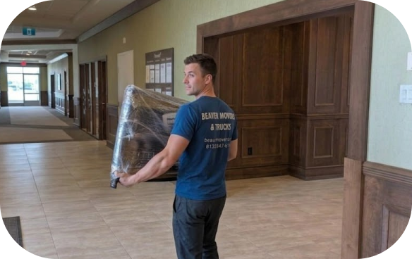 Beaver Movers & Trucks mover in a blue company shirt carrying an item fully wrapped in clear shrink wrap through a bright hallway with tile floors