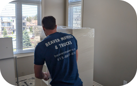 Beaver Movers & Trucks crew member applying clear shrink wrap to a tall white cabinet in a bright home