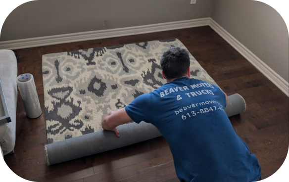 A Beaver Movers professional from behind, kneeling on a dark wood floor and rolling up a large patterned area rug, with a roll of clear stretch wrap nearby and Beaver Movers branding on his blue shirt